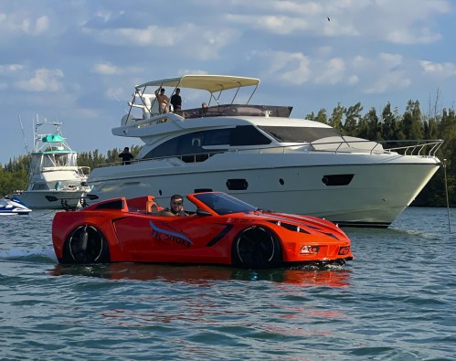 A bright orange sports car boat cruising in the water, with a luxury yacht in the background and people on board.