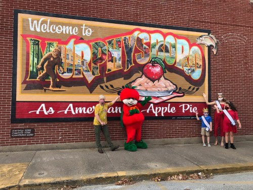 A colorful mural welcomes visitors to Murphysboro, featuring a mascot and people posing in front of it.
