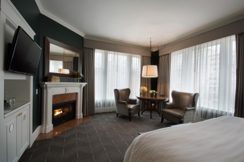 Cozy hotel room featuring a fireplace, two chairs, a round table, and large windows with soft curtains.