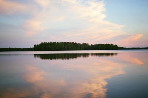 A serene lake at sunset, reflecting soft clouds and trees on the horizon, creating a peaceful atmosphere.