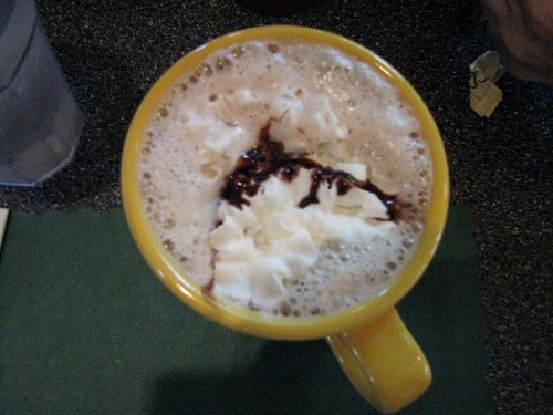 A yellow mug filled with frothy coffee topped with whipped cream and chocolate drizzle.