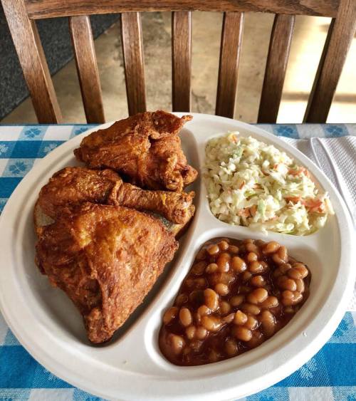 A plate with three pieces of fried chicken, coleslaw, and baked beans, set on a checkered tablecloth.