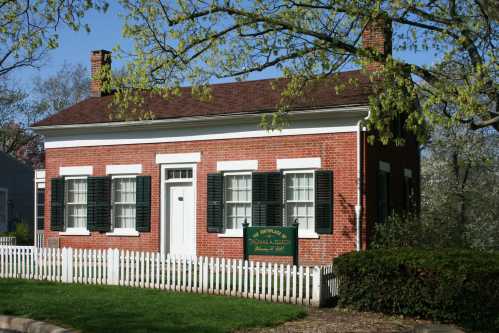 A small red brick house with white trim, green shutters, and a picket fence, surrounded by trees and grass.