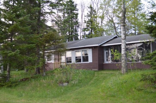 A brown house surrounded by trees, featuring a porch and large windows, set in a grassy area.