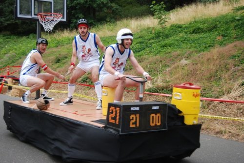 Three men in sports attire ride a makeshift basketball cart, with a scoreboard displaying 21-0, on a sloped track.
