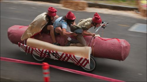 Three people in costumes riding a large, inflatable hot dog on a bicycle during a fun event.