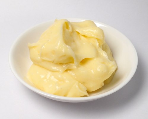 A small white bowl filled with creamy, smooth yellow mayonnaise.