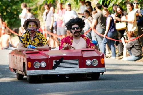 Two people in colorful outfits drive a homemade go-kart down a street, surrounded by spectators.