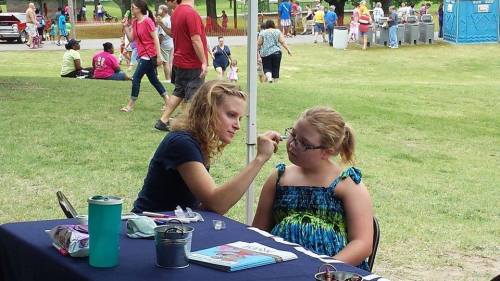 A girl sits while an artist paints her face at an outdoor event, with people walking in the background.