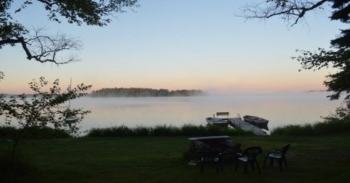 Serene lakeside view at dawn, with mist rising over the water and boats docked near a grassy shore.