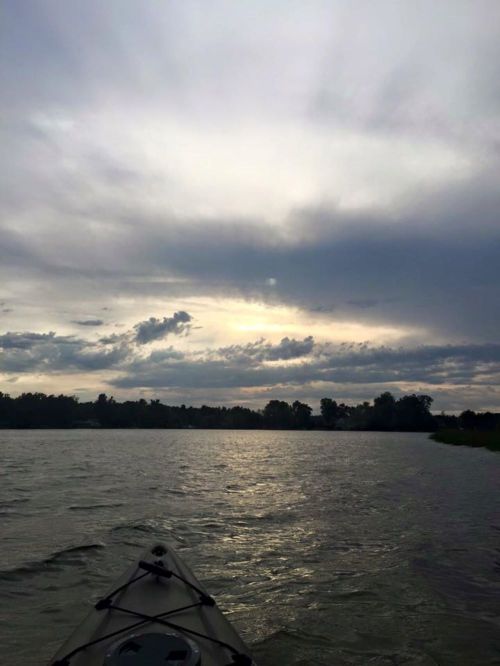 A kayak on calm water under a cloudy sky, with the sun peeking through the clouds in the distance.
