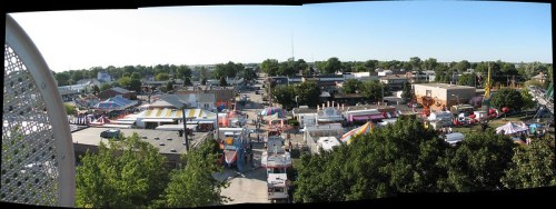 A panoramic view of a bustling fairground with colorful tents, rides, and a clear blue sky.