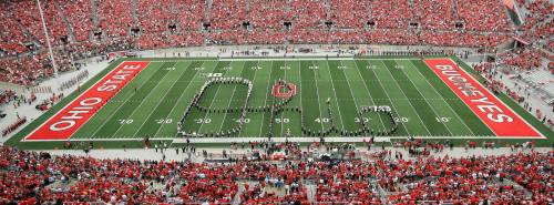 Aerial view of a packed stadium with fans in red, featuring a marching band forming "BAND" on the field.