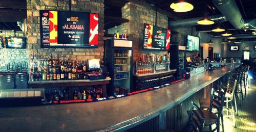 A spacious bar with a long counter, shelves of drinks, and large screens displaying events in a modern setting.