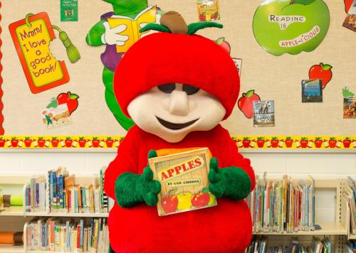 A mascot dressed as an apple holds a book titled "Apples" in a colorful library setting with apple-themed decorations.