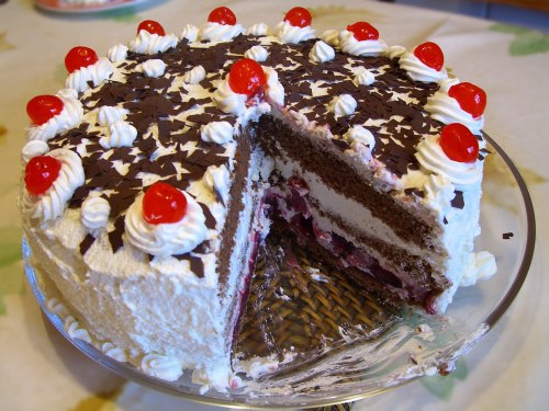 A chocolate cake with whipped cream, cherries on top, and a slice cut out, displayed on a glass cake stand.