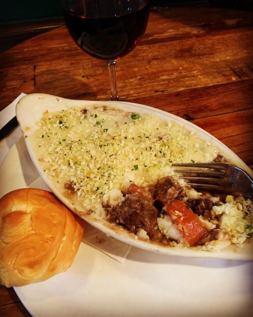 A dish of baked meat with vegetables, topped with cheese, served with a roll and a glass of red wine.