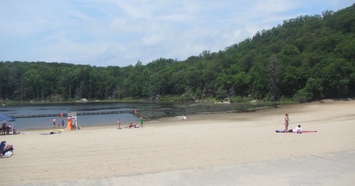A sandy beach by a calm lake, surrounded by green trees, with people relaxing and enjoying the sun.