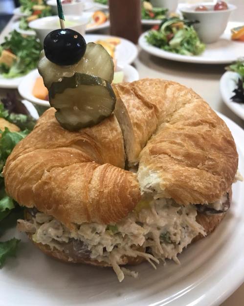 A croissant sandwich filled with chicken salad, topped with a pickle and olive, served with a side of salad.