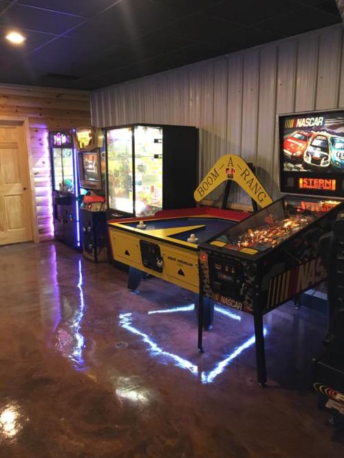 A game room featuring arcade machines, a pinball machine, and a colorful air hockey table against a wooden wall.