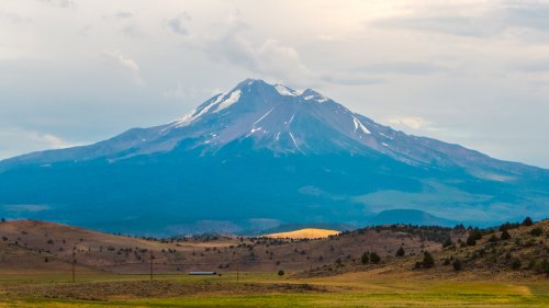 A majestic mountain with snow-capped peaks rises above a grassy landscape under a cloudy sky.