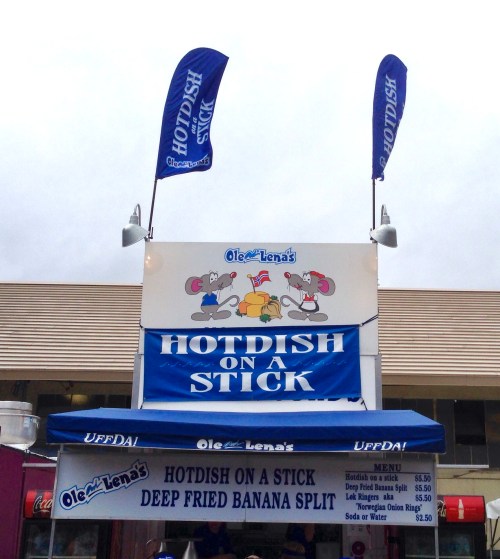 A food stand with a sign reading "Hotdish on a Stick" and a menu featuring deep-fried banana split. Flags above.