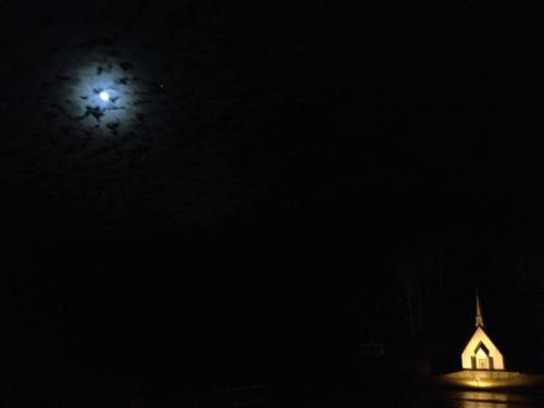 A small illuminated chapel stands in darkness, with a bright moon partially obscured by clouds in the night sky.