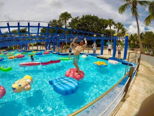 A person jumps on colorful floating toys in a pool, surrounded by palm trees and other people enjoying the water.