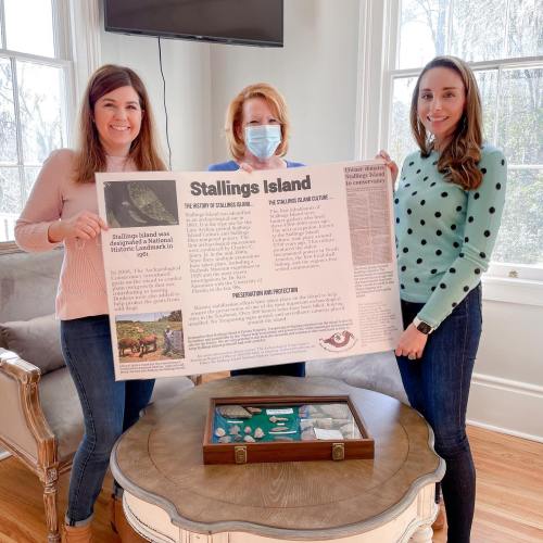 Three women hold a large poster about Stallings Island, with artifacts displayed on a table in front of them.