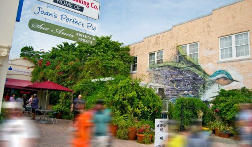 A colorful mural of a bird on a building surrounded by greenery, with people walking by and signs for local businesses.