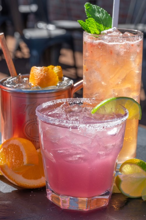 Three colorful cocktails on a table, garnished with fruit and mint, with fresh citrus slices nearby.