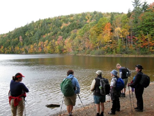 A group of hikers stands by a lake, surrounded by colorful autumn foliage on the hills in the background.