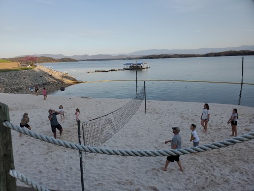 A group of people playing beach volleyball on a sandy shore by a calm lake with mountains in the background.