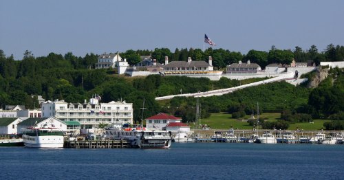 A scenic view of a waterfront with boats, lush greenery, and a hilltop featuring buildings and an American flag.