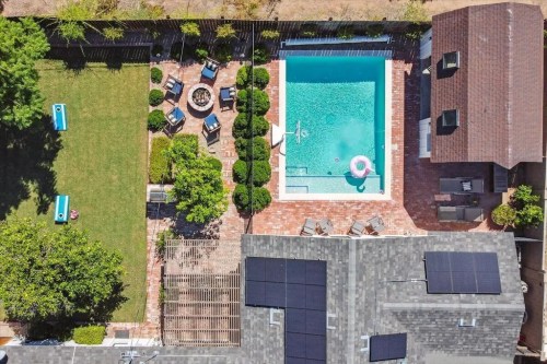 Aerial view of a backyard with a pool, lounge chairs, a fire pit, and landscaped greenery. Solar panels on rooftops.