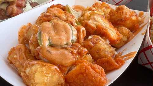 A serving of crispy fried cauliflower bites drizzled with spicy sauce, served in a white paper tray.