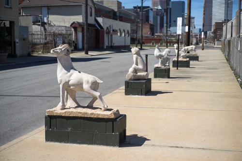 A row of stone sculptures depicting animals and figures along a city sidewalk on a sunny day.