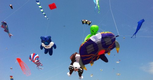 Colorful kites in various shapes, including a bear and an alien, flying against a clear blue sky.