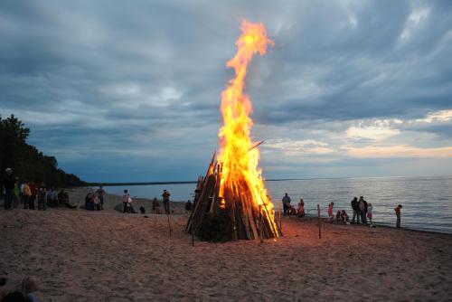 A large bonfire on a beach at dusk, with people gathered around and a calm lake in the background under cloudy skies.