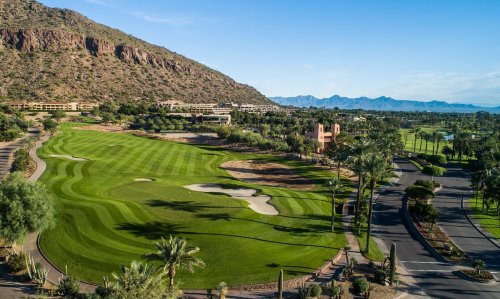 Aerial view of a lush golf course surrounded by mountains and palm trees under a clear blue sky.
