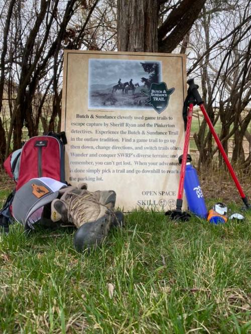 A backpack and hiking poles rest on grass near a sign about the Butch & Sundance trail and its history.