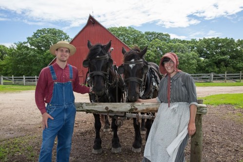 A man and woman in traditional attire stand beside two horses in front of a red barn on a sunny day.