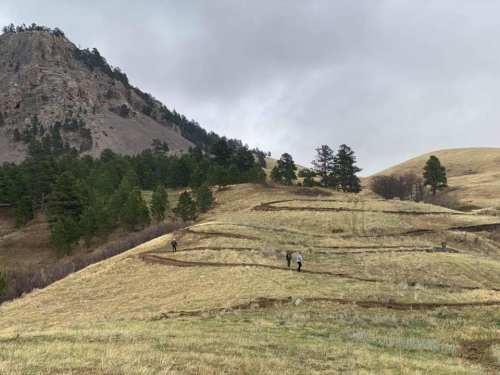 Two hikers walk along a winding trail in a grassy landscape with rolling hills and scattered trees under a cloudy sky.