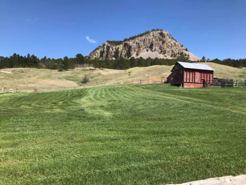 A green field with a red barn in the foreground and a rocky mountain in the background under a clear blue sky.