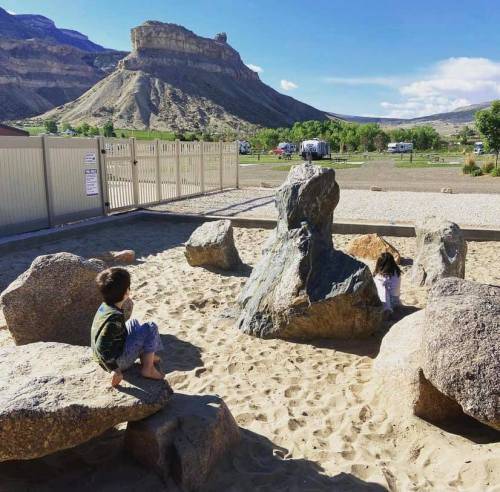 Two children play on large rocks in a sandy area, with a mountainous landscape and blue sky in the background.