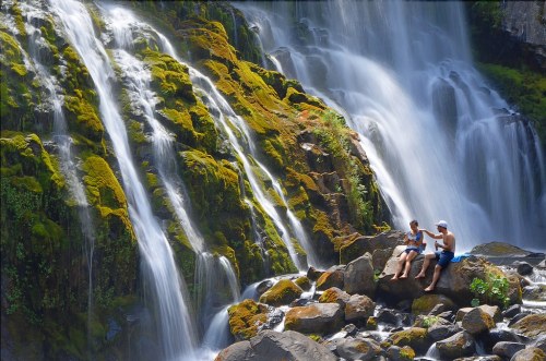 Two people sit on a rock by a waterfall, surrounded by lush greenery and flowing water.