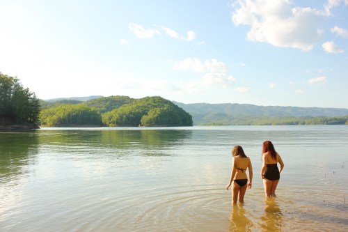 Two women wade into a calm lake surrounded by green hills and a clear blue sky.