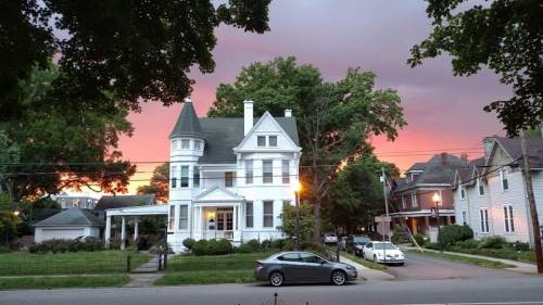 A large white Victorian house with a turret, surrounded by trees, under a colorful sunset sky.