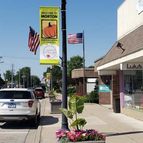 Street view of Morton, featuring a welcome banner, American flags, and colorful flower planters along the sidewalk.