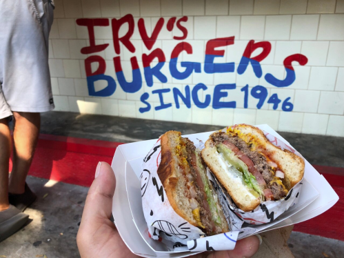 A hand holding a burger with layers of meat and toppings, in front of a colorful mural reading "Irv's Burgers Since 1946."